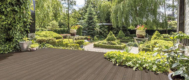 Eine schicke Terrasse aus dunklem Holz. Im Hintergrund sieht man einen gepflegten Garten mit vielen Pflanzen und Bäumen.